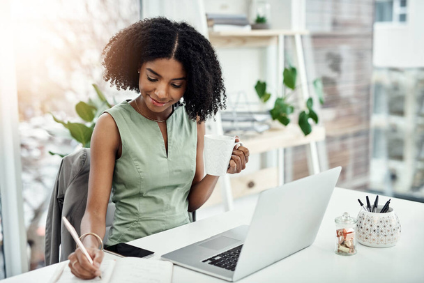 Black woman, writing and office research at laptop as journalist with notebook, creative or project planning. Female person, smile and coffee up in startup agency for brief, review or brainstorming. - Photo, Image