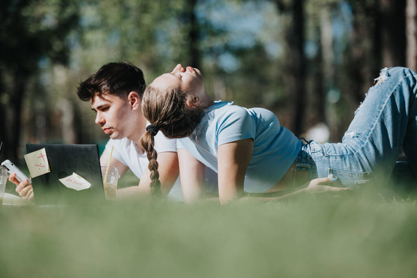 A school couple lies back-to-back outdoors, immersed in their own activities, sharing a peaceful moment together, symbolizing youthful companionship. - Photo, Image