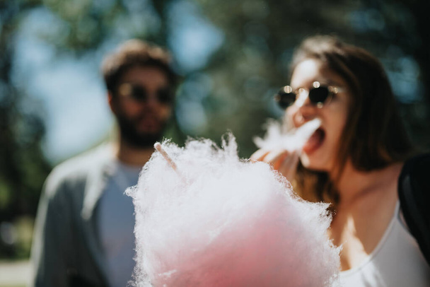 Веселая молодая женщина в солнечных очках наслаждается вкусом розовой сахарной ваты, разделяя радостный момент под летним солнцем, в то время как размытый мужчина наблюдает на заднем плане. - Фото, изображение