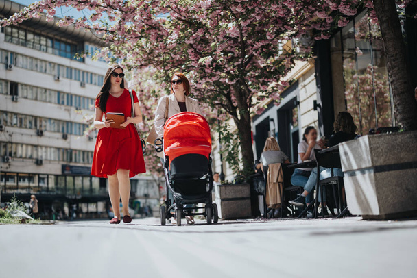 Empresarias vestidas profesionalmente pasean con un cochecito rojo entre árboles florecientes en un entorno urbano. - Foto, imagen