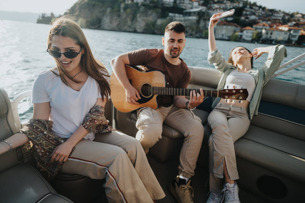Молоді дорослі розслабляються і розважаються на човні, коли грають на гітарі, у супроводі мальовничих краєвидів і літніх вітрів. - Фото, зображення