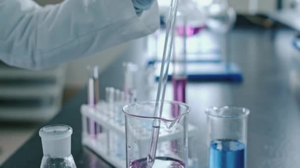 Close-up beelden van handschoenen van onherkenbare laboratoriumtechnicus die lange glazen pipet vullen met roze chemisch reagens, toevoegen aan heldere vloeistof in kolf, dan roeren in cirkelvormige beweging - Video