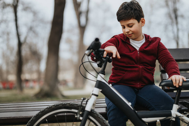 Eine ruhige Szene fängt eine junge Person ein, die auf einer Bank neben ihrem Fahrrad innehält und einen friedlichen Moment in einer grünen Parkumgebung genießt.. - Foto, Bild