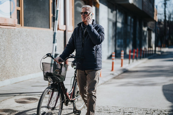 Un adulto maduro activo charla en su teléfono mientras está de pie con su bicicleta en una acera urbana, mostrando conectividad moderna y estilo de vida. - Foto, Imagen
