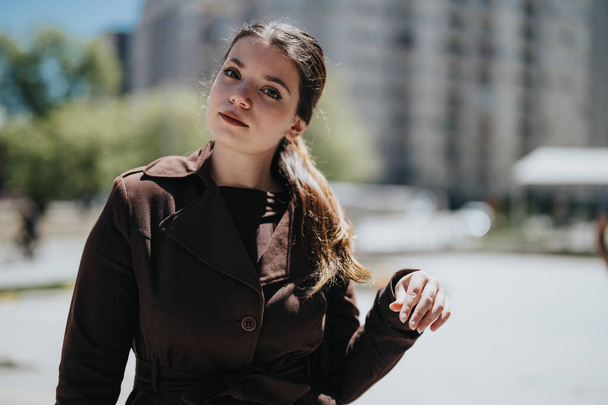 Una joven profesional se encuentra confiada en un parque urbano, vistiendo un abrigo marrón de moda, exudando confianza y estilo. - Foto, Imagen