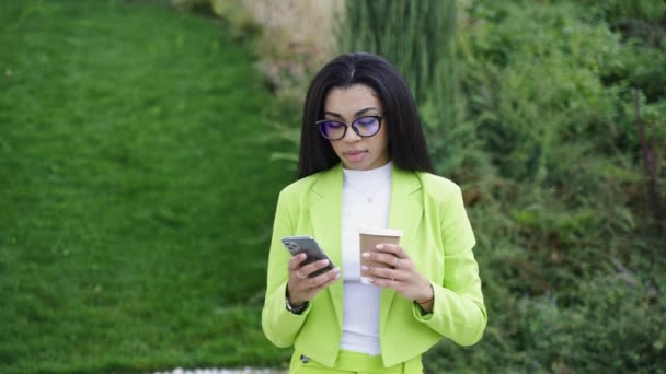Γυναίκα με πράσινα ρούχα γραφείου, κρατώντας smartphone, στέκεται στο πάρκο, και πίνοντας καφέ. Αργή κίνηση - Πλάνα, βίντεο