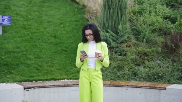 Mujer vestida con ropa de oficina verde, sosteniendo un smartphone, parada en el parque y bebiendo café. Movimiento lento - Imágenes, Vídeo