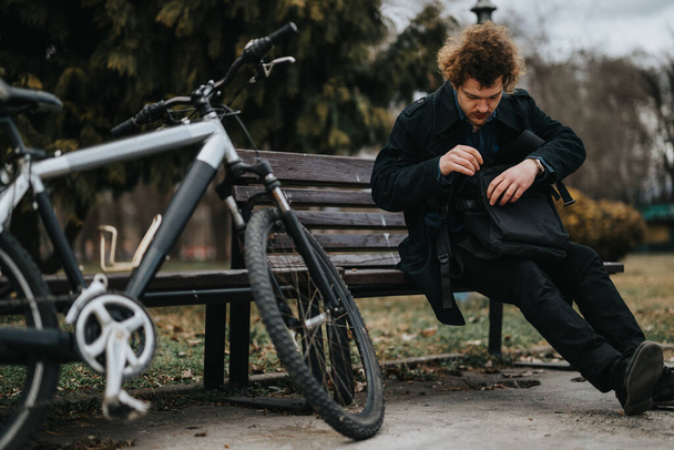 Ein ehrgeiziger junger Geschäftsmann erledigt die Arbeit aus der Ferne am Laptop, während er mit dem Fahrrad auf einer Parkbank sitzt. - Foto, Bild
