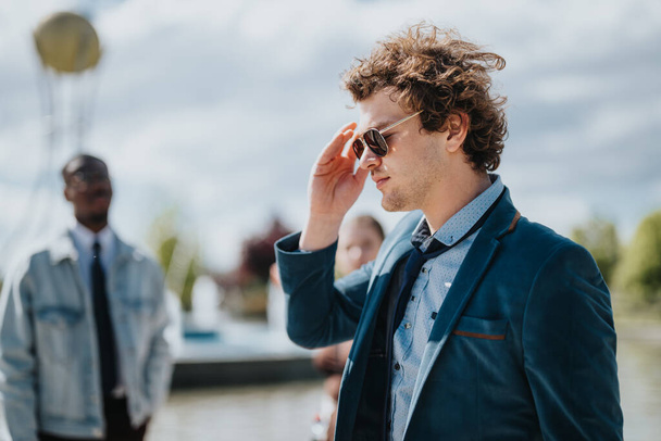 A stylish young businessperson adjusting his sunglasses stands confidently outdoors. In the blurry background, colleagues are visible. - Photo, Image