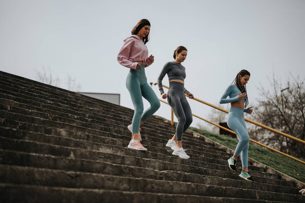 Három tornafelszerelésben lévő atlétikai nő ereszkedik le egy hosszú lépcsőn, miközben városi szabadtéri környezetben edzenek, elősegítve az egészséget és a fitnessz állapotot.. - Fotó, kép