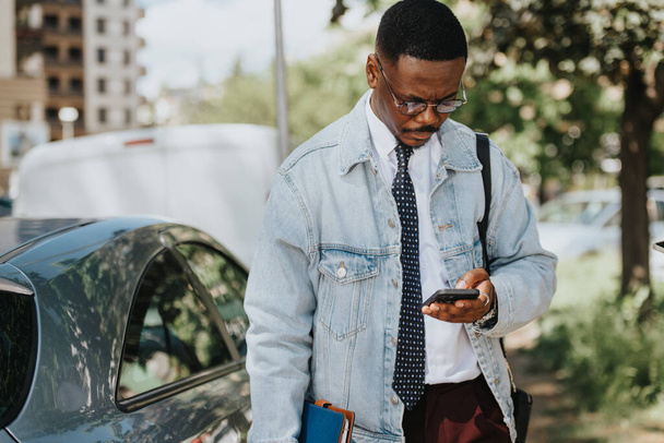 Професійний афро-американський бізнесмен в стильній джинсовій куртці, використовуючи свій смартфон поруч зі своєю машиною в міському середовищі в сонячний день. - Фото, зображення