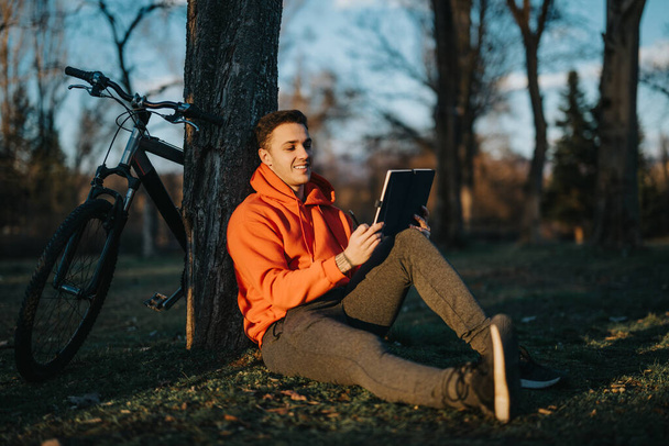 Zwyczajny młody człowiek cieszący się wolnym czasem w przyrodzie, czytając na notatniku z rowerem opartym na drzewie. - Zdjęcie, obraz