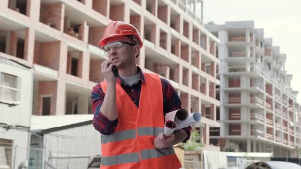 Builder with rolled-up architectural drawings under his arm talking on walkie-talkie while standing among unfinished multi-story buildings. Slow motion - Footage, Video