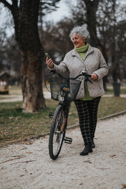 Una mujer madura con el pelo rizado gris de pie con su bicicleta en un parque, sosteniendo un teléfono inteligente. - Foto, imagen