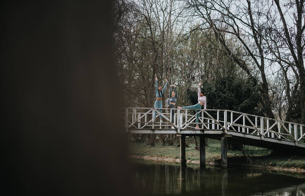 Three women in athletic wear stretching on a bridge in a serene park, embodying health and fitness. - Photo, Image