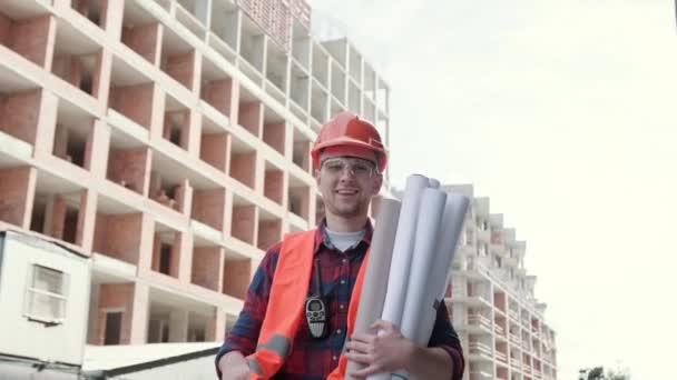 Porträt eines gut gelaunten Poliers mit Bauplänen, der zwischen unfertigen mehrstöckigen Gebäuden steht und vor der Kamera ein OK-Zeichen setzt. Zeitlupe - Filmmaterial, Video