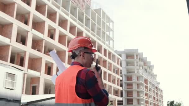 Achteraanzicht van de bouwmanager met tekeningen die praten op walkietalkie terwijl hij voor onafgewerkte huizen met meerdere verdiepingen staat. Langzame beweging - Video