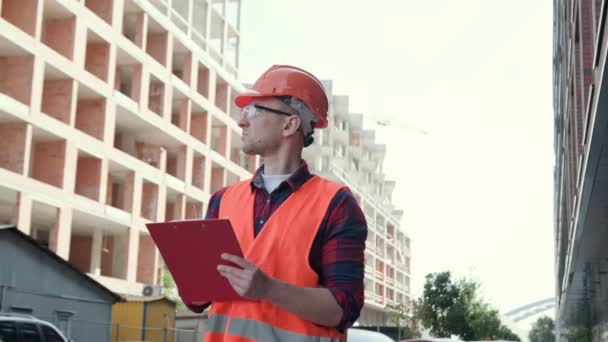 Serieuze bouwinspecteur schrijft iets op klembord terwijl hij onafgewerkt huis met meerdere verdiepingen inspecteert. Langzame beweging - Video