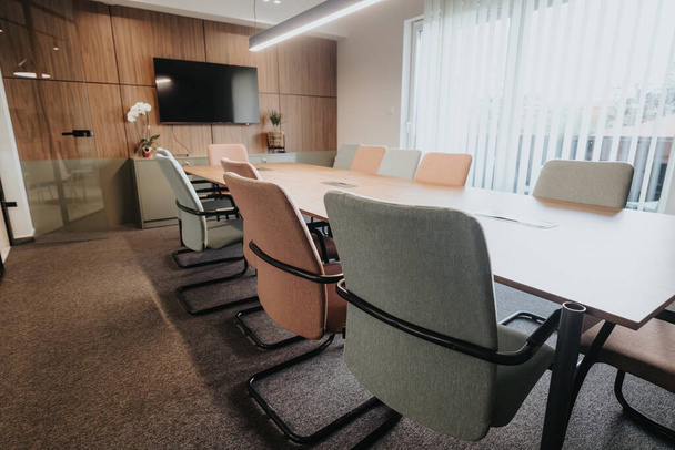 Deze moderne kantoorvergaderruimte beschikt over een houten muur, een grote tafel, stijlvolle stoelen en een gemonteerde tv, ideaal voor zakelijke bijeenkomsten.. - Foto, afbeelding