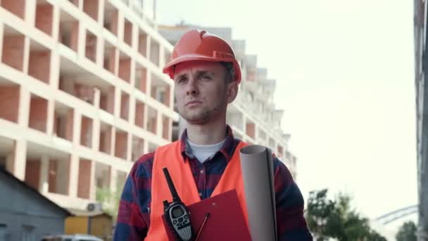 Portrait of serious foreman with walkie-talkie and drawings looking into distance while standing between unfinished multi-story buildings. Slow motion - Footage, Video
