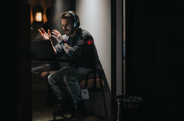 Hombre con auriculares haciendo gestos mientras juega por la noche, mostrando emoción y enfoque, en una habitación con luz de mal humor. - Foto, Imagen