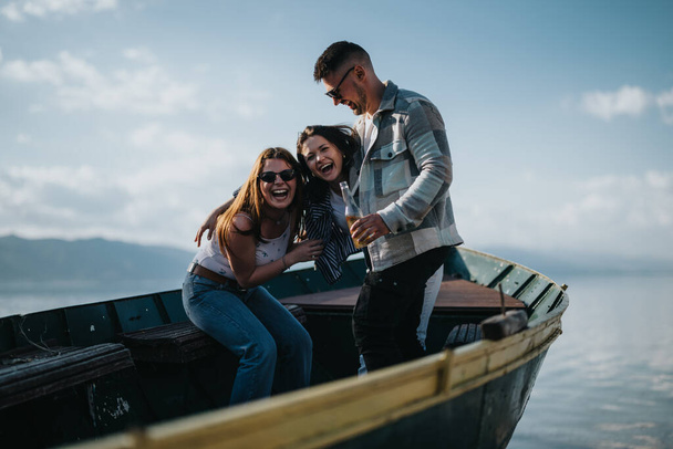 Három barátból álló csoport együtt nevetett és ivott egy hajón, egy festői tóval és hegyekkel a háttérben egy napsütéses napon.. - Fotó, kép