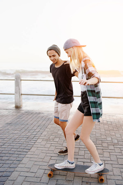 Paar, man en vrouw met skateboard op de promenade, gelukkig en behulpzaam voor evenwicht, leren en speels over zee. Mensen, lesgeven en schaatsen met ondersteuning en plezier op de boulevard voor vakantie in Italië. - Foto, afbeelding