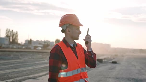 Foreman en ropa protectora y casco sosteniendo walkie-talkie y hablando con los constructores, da instrucciones. Movimiento lento - Metraje, vídeo