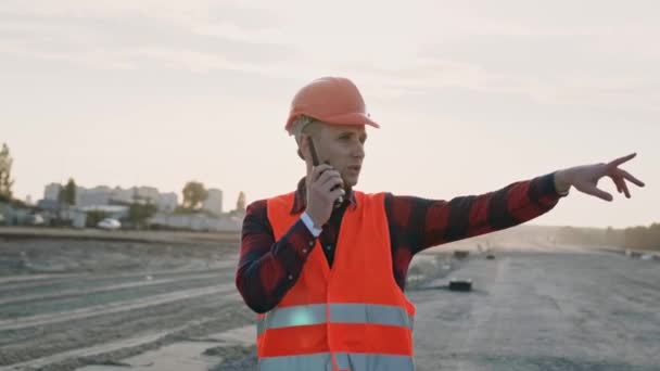 El hombre con ropa protectora y casco sosteniendo walkie-talkie y hablando con los constructores, da instrucciones. Movimiento lento - Metraje, vídeo