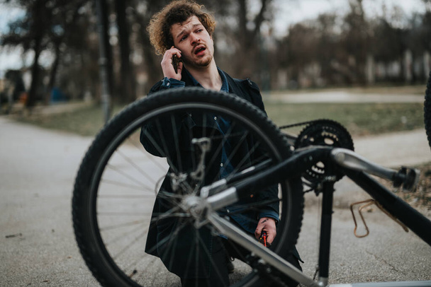 Een jonge mannelijke ondernemer is multitasking door het repareren van zijn fiets en het spreken aan de telefoon in een park instelling. - Foto, afbeelding