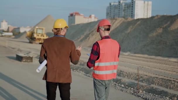 Professional foreman in protective clothes and helmet walking with customer on site, excavator passes nearby. Slow motion - Footage, Video