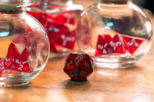 A single red die stands prominently in focus, contrasting with blurred dice in glass jars in the background, on a rustic wooden table - Photo, Image