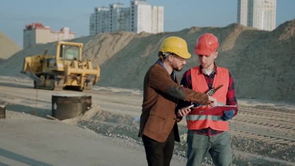 Foreman en ropa protectora y casco caminando con el cliente en el sitio, excavadora pasa cerca. Movimiento lento - Metraje, vídeo