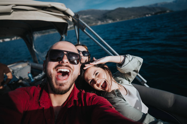 Группа друзей, разделяющих смех и наслаждение на лодке, делающих игривое селфи на фоне видов на озеро во время солнечного отпуска - Фото, изображение