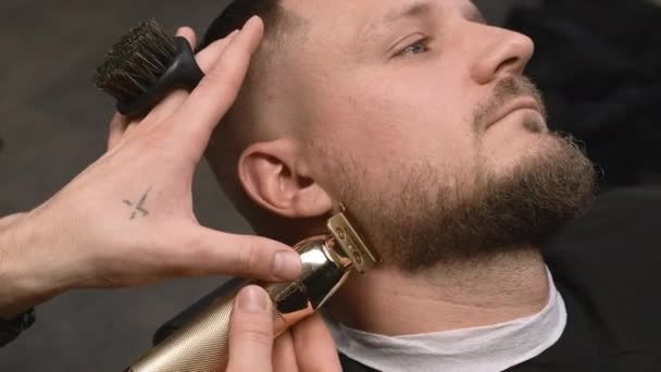 Hairstylist faz forma de barba elegante com aparador para o cliente em barbearia closeup. Barbeiro corta o cabelo facial do homem usando a máquina no salão - Filmagem, Vídeo