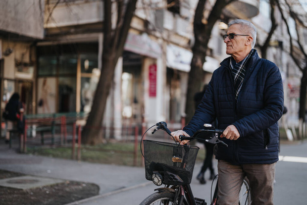 Kypsä mies, jolla on polkupyörä, seisoo kaupunkien jalkakäytävällä, mikä heijastaa rentoa elämäntapaa ja aktiivista ikääntymistä kaupunkiympäristössä.. - Valokuva, kuva