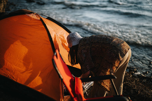 Femme se penchant alors qu'elle installe une tente orange vif au bord du lac pittoresque, capturant un moment de détente et de plaisir en plein air avec des amis. - Photo, image