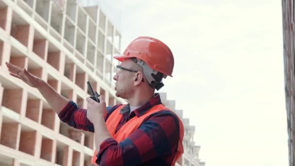 Egy férfi védőruhában és sisakban az épület mellett áll, walkie-talkie-val a kezében és munkásokkal beszél. Lassú mozgás. - Felvétel, videó