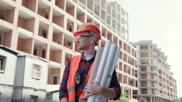 Constructor en ropa protectora y casco de pie cerca del edificio, sosteniendo planos, mostrando señal de ok. Movimiento lento - Metraje, vídeo