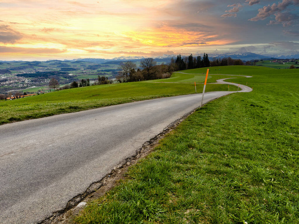 Impresionante puesta de sol sobre un exuberante paisaje suizo, con un sinuoso camino a través de campos verdes vibrantes que conducen a los majestuosos Alpes nevados, bajo un cielo dramático pintado con tonos de naranja y azul - Foto, Imagen