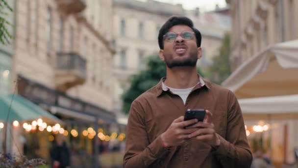 Onneton Intian etninen mies matkustaja mies kaveri turisti tutkia kaupungin reitti matkustaa ulkona. Liikemies kävellä ulkomailla katu näköinen matkapuhelin gadget matkapuhelin kävellä kaupunki käyttäen online-kartta - Materiaali, video