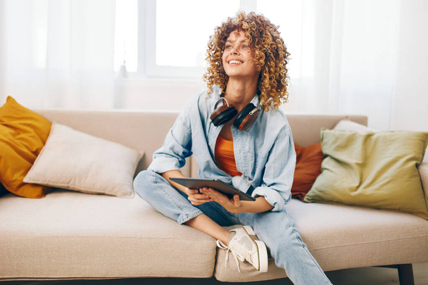 Szczęśliwa kobieta trzyma tablet PC, słuchając muzyki ze słuchawkami, relaksując się na kanapie w domu - Zdjęcie, obraz