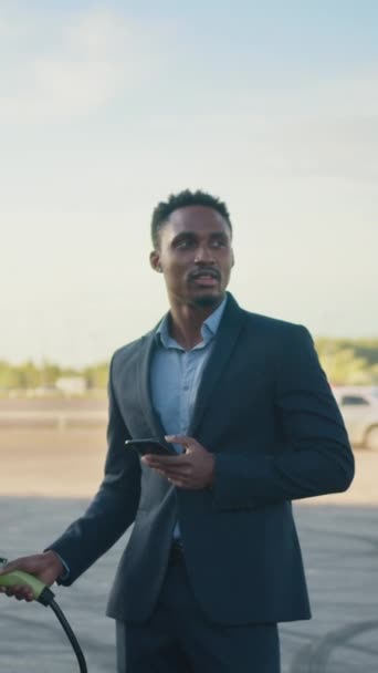 垂直スクリーン:電気自動車の充電からケーブルで屋外に立っている間現代スマートフォンを使用してスーツのアフリカのアメリカのビジネスマンの閉鎖. 現代技術,ビジネス,コンセプト - 映像、動画
