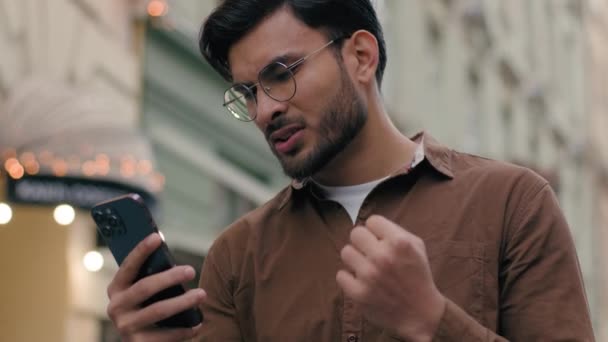 Lähikuva järkyttynyt onneton intialainen etninen mies nuori kaveri liikemies mies etsii matkapuhelin huono yhteys menettää onni menetys epäonnistua kosketus pään miksi negatiivinen tulos ulkona kaupunki kaupunkien katu - Materiaali, video