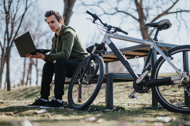 Przystojny młody człowiek używa laptopa siedząc na ławce w parku, z zaparkowanym obok rowerem, w pogodny, słoneczny dzień. - Zdjęcie, obraz