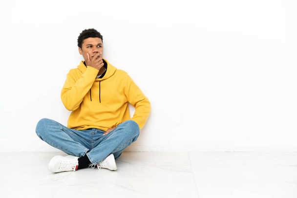 Молодий афроамериканець, що сидить на підлозі ізольований на білому фоні, має сумніви і плутають вираз обличчя. - Фото, зображення