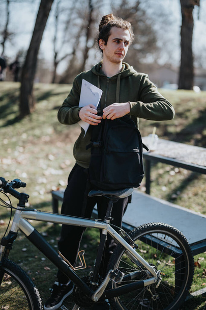 Un joven reflexivo se relaja con su bicicleta de montaña en un parque iluminado por el sol, agarrando un cuaderno, exudando una sensación de calma y contemplación en medio de la naturaleza. - Foto, Imagen