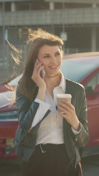Ekran pionowy: Kobieta wykonująca rozmowę wideo na smartfonie podczas ładowania samochodu. Urocza biała kobieta, która podczas ładowania luksusowego elektrycznego samochodu rozmawia przez nowoczesny smartfon. Pojęcie ludzi - Materiał filmowy, wideo