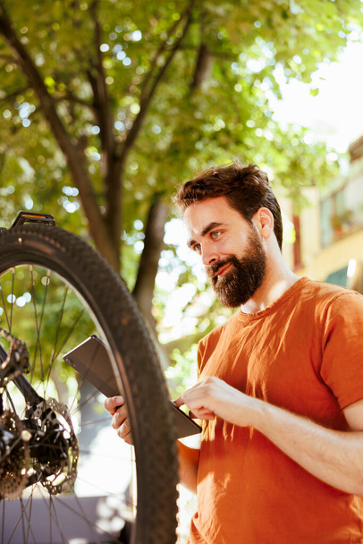 Entuzjastyczny zdrowy rowerzysta sprawdzanie opon jako coroczny rutyna konserwacji gumy rowerowej z tabletu cyfrowego. Kaukaski mężczyzna za pomocą inteligentnego urządzenia do poszukiwania rozwiązań uszkodzonych opon rowerowych. - Zdjęcie, obraz