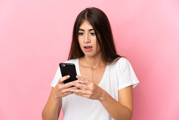 Young caucasian woman isolated on pink background looking at the camera while using the mobile with surprised expression - Photo, Image